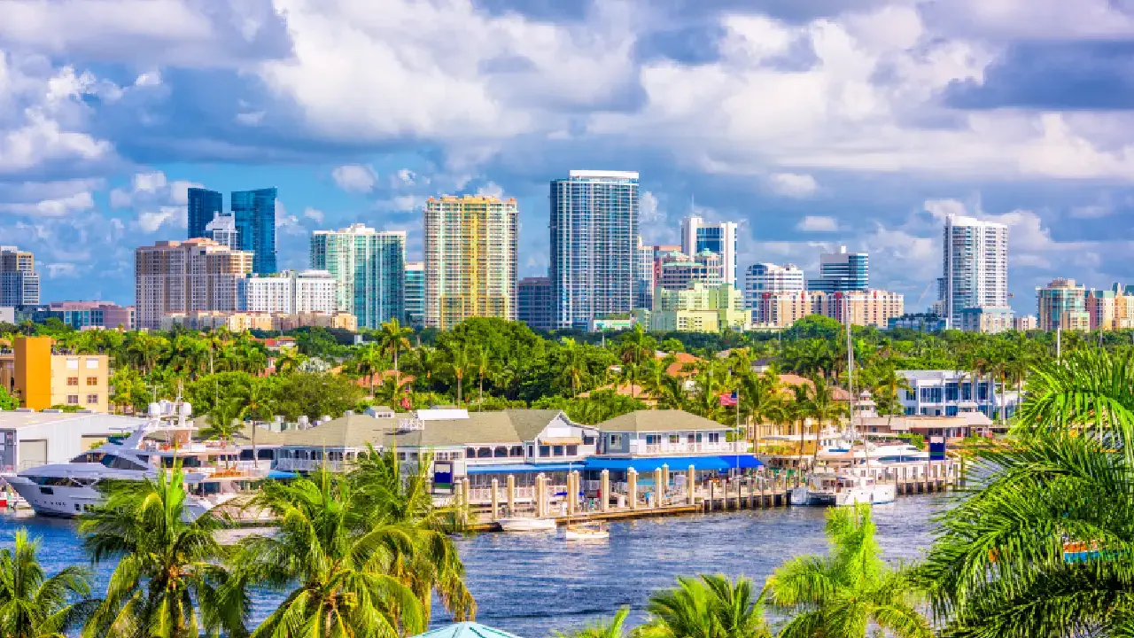 a vibrant Florida cityscape with towering modern high-rises against a bright blue sky filled with fluffy clouds. In the foreground, lush green palm trees and waterfront buildings line a marina, complete with yachts and boats docked along the calm water. A tropical, colorful urban paradise.