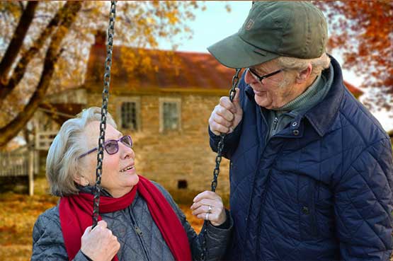 An older couple joyfully swings together in a park surrounded by colorful autumn leaves.