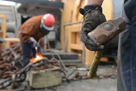 A man wearing protective gear welds two pieces of metal together in a workshop setting. Sparks fly from the welding process.
